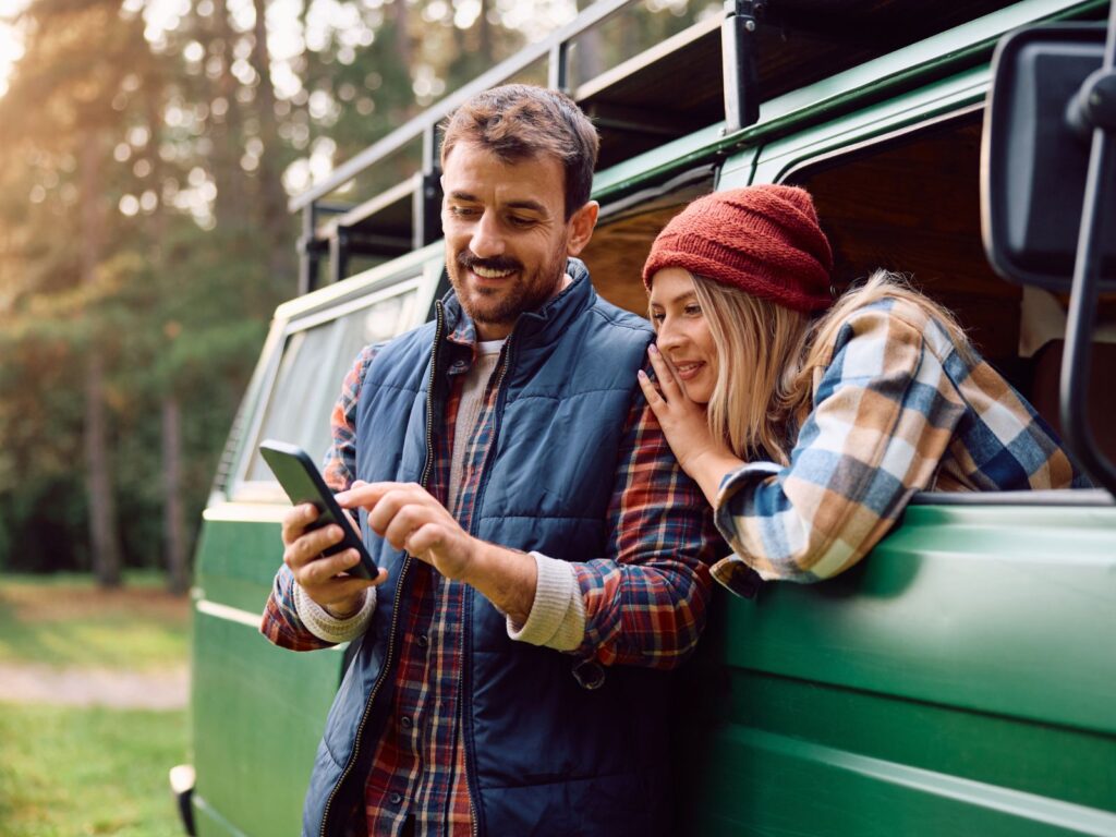 Happy couple using smartphone outside campervan in forest