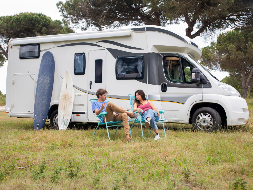 Young couple (man and woman) sat on camping chairs in a field, next to their campervan and a surfboard 